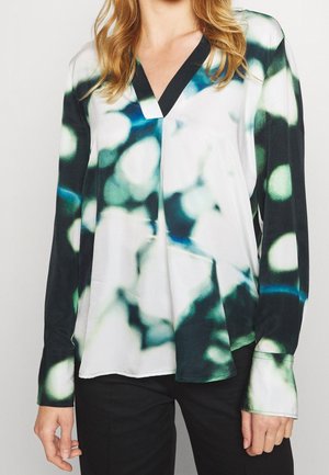 Femme portant une blouse à manches longues avec un motif abstrait noir, blanc et vert et un col en V, associée à un pantalon noir.