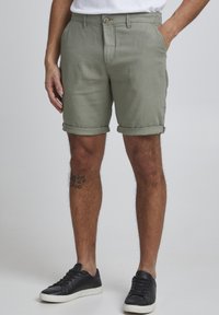 Pantalones cortos de color verde oliva claro hechos de una tela texturizada, con cierre de botón, dobladillos enrollados y bolsillos laterales. Zapatillas negras con suelas blancas.
