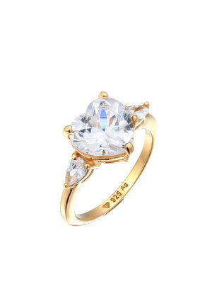 Guldbelagt ring med en stor hjerteformet klar ædelsten i midten, flankeret af to mindre pæreformede sten, mærket "925 AG."