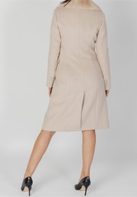 Abrigo de mezcla de lana beige con un cuello de piel sintética, ajuste entallado, mangas largas y detalles de botones en los puños. Combinado con tacones altos negros.