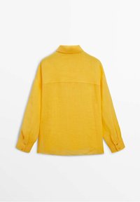 Chemise en lin jaune à manches longues avec col, dotée d'un empiècement au dos et de poignets boutonnés. Le tissu paraît léger et texturé.