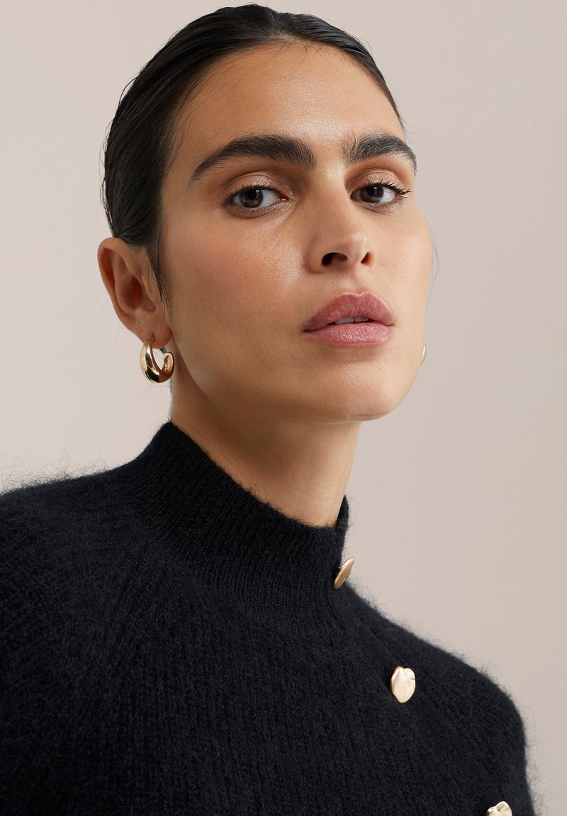 Vrouw met slicked-back haar draagt gouden creolen en een zwarte gebreide trui versierd met glanzende gouden knopen op een neutrale achtergrond.