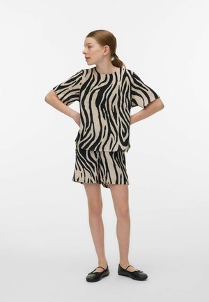 Kvinde med lyst brunt hår i en hestehale iført en kortærmet sort og beige zebra-print top og matchende shorts, stående med hænderne på hofterne.