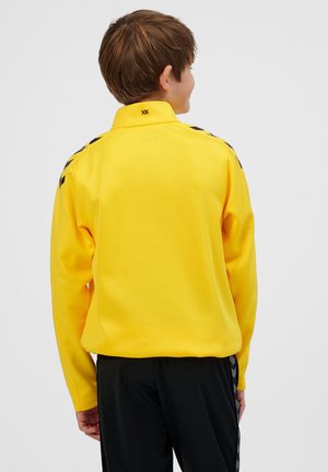 Gelbe Jacke mit hohem Kragen, langen Ärmeln und schwarzen Akzenten an den Schultern. Glatte Textur, lockere Passform und ein Logo nahe dem Kragen.
