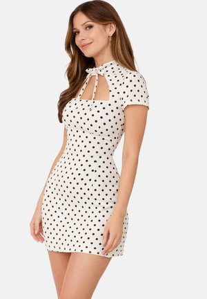 Femme portant une mini-robe blanche ajustée à manches courtes avec des pois noirs et un décolleté en trou de serrure noué avec un nœud.