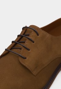 Καφέ δερμάτινο παπούτσι τύπου δερμπέρι από σουέντ με στρογγυλά σκουροκαφέ κορδόνια και ορατές ραφές στις θηλιές και τις άκρες.