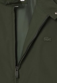 Giacca verde scuro realizzata in tessuto liscio con chiusura a zip e logo ricamato sul petto. Presenta bottoni a pressione al colletto.