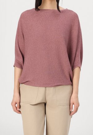 Vrouw draagt een losse mauve geribbelde gebreide top met ellebooglange mouwen en beige broek met voorzakken.