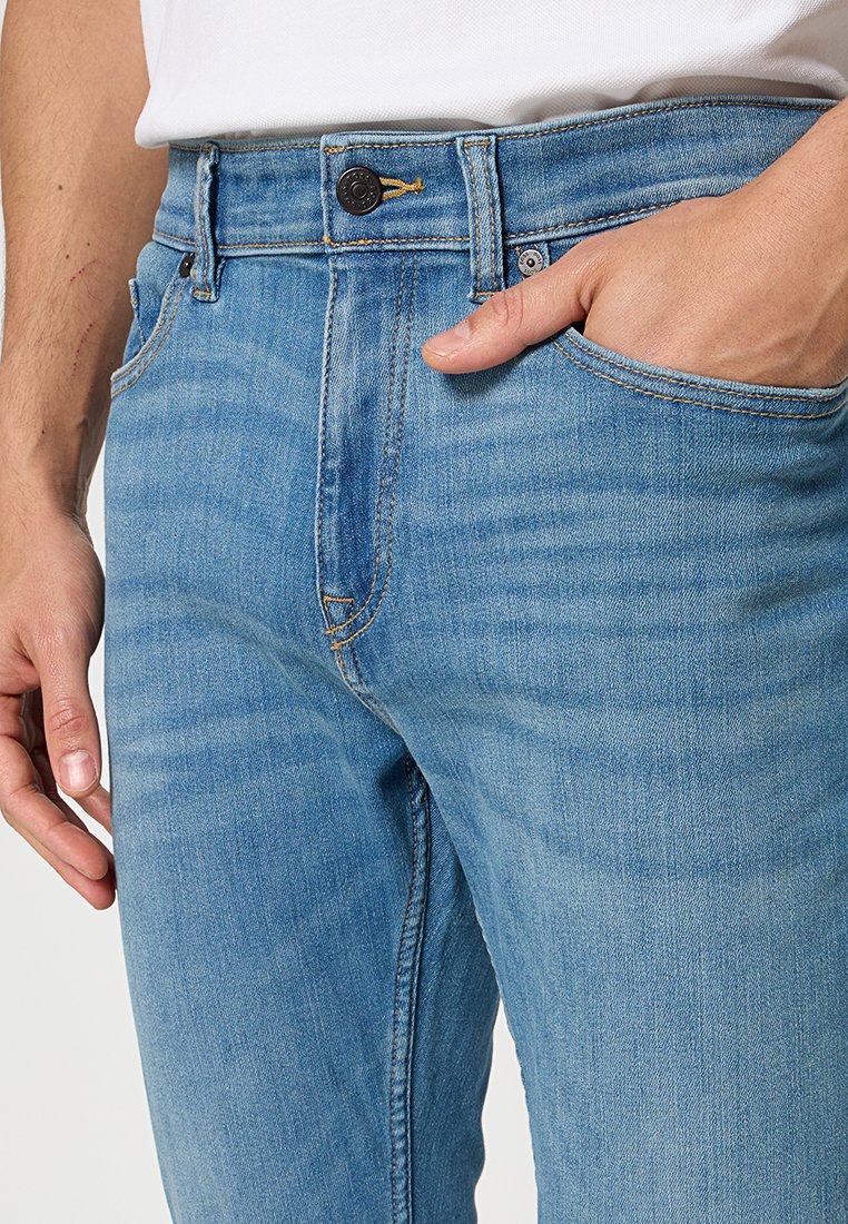 Jean en denim bleu clair avec une coupe slim, présentant une poche avant visible et des accents de couture marron. Fermeture à bouton avec un détail de fil jaune.