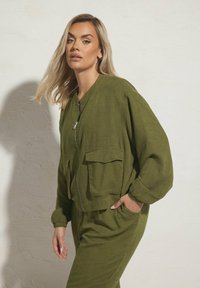 Giacca bomber verde con zip frontale, due tasche anteriori, polsini arricciati e vestibilità rilassata. Realizzata in tessuto texturizzato, abbinata a pantaloni coordinati.