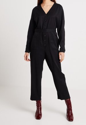 Frau in einem schwarzen Langarm-Jumpsuit mit Knöpfen und Fronttaschen sowie bordeauxroten, hochhackigen Stiefeletten vor einem schlichten Hintergrund.
