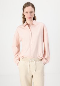Light pink button-up shirt with long sleeves and cuff detailing, paired with beige sweatpants featuring a drawstring and subtle logo.