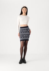 Jupe tricotée en navy avec un motif Fair Isle blanc, présentant des rennes et des flocons de neige. Associée à un haut ajusté à manches longues et des chaussures noires.