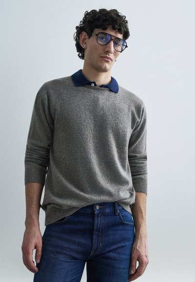 Grå strikket sweater med navy krave, der har lange ærmer og ribbede manchetter, kombineret med mørkeblå jeans. Runde briller med metalstel.