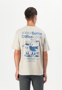 Beige Baumwoll-T-Shirt mit blauer Grafik von zwei Cartoon-Charakteren, die einem Glas und einer Kaffeekanne ähneln, mit Text und Adresse darunter.