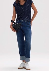 Laadukkaasta tummansinisestä lyhythihaisesta paidasta, jossa on valkoisia pilkkuja, vaaleansinisistä käänteistä farkuista ja valkoisista tennareista, joissa on beigeä korostetta; kädessä pieni musta laukku.