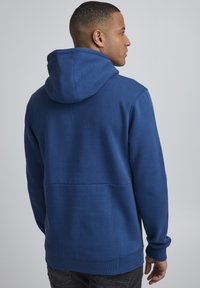 Sudadera con capucha azul en una tela suave, con un bolsillo tipo canguro y puños de canalé, que presenta un corte holgado y un diseño sencillo.