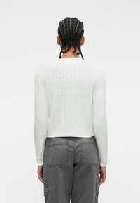 Biely svetrový pulóver s dlhými rukávmi a skráteným strihom, skombinovaný so šedými cargo nohavicami s dvojitými zadnými vreckami.