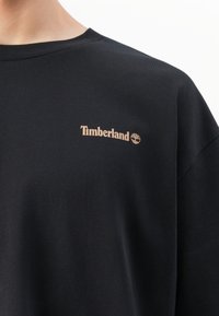 Gros plan sur un t-shirt noir à col rond avec un petit logo beige de Timberland sur le côté gauche de la poitrine.
