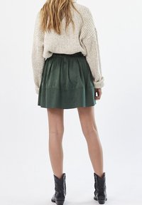 Mini jupe en simili cuir vert avec des plis et une taille élastique, associée à un pull oversize en tricot crème et des bottines noires.