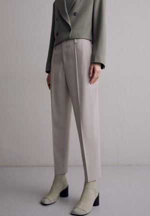 Olijfgroene dubbelzijdige blazer gedragen met beige op maat gemaakte broeken, geaccentueerd door lichtgroene hoge hakken en bijpassende sokken.