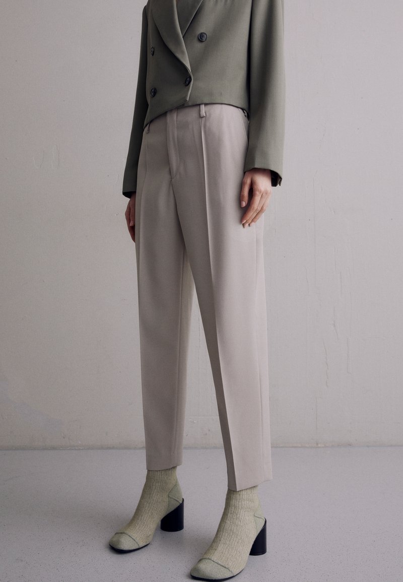 Olivově zelený dvouřadý blejzr v kombinaci s béžovými upravenými kalhotami, doplněný světle zelenými lodičkami na vysokém podpatku a odpovídajícími ponožkami.