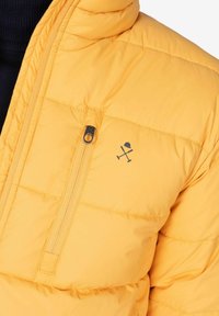 Chaqueta de plumas amarilla con diseño acolchado, que cuenta con un bolsillo con cremallera y un logo de herramientas cruzadas. Textura suave y cuello con capucha.