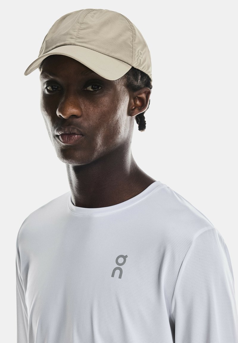 Cappello beige con visiera curva, realizzato in tessuto leggero. Camicia bianca a maniche lunghe con un sottile motivo testurizzato e un logo grigio.