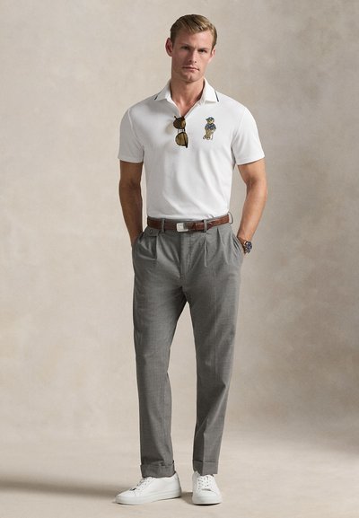 Λευκό πόλο μπλουζάκι με σκουρόχρωμη λεπτομέρεια στο γιακά και κεντημένο σχέδιο, σε συνδυασμό με ανοιχτό γκρι πλισέ παντελόνι και λευκά αθλητικά παπούτσια.