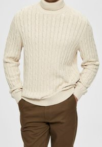 Crème-kleurige kabelgebreide trui met hoge kraag, geribde mouwen en zoom. Draagt over een bruine broek, die textuurdetails laat zien.