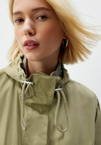 Khaki bunda s kapucňou a sťahovacou šnúrkou, obsahujúca strieborné kovanie a hladkú textúru. Model má svetlo blond vlasy.