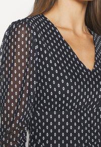 Schwarzes gemustertes Kleid mit V-Ausschnitt, durchsichtigen langen Ärmeln, weißen Rauten und strukturiertem Stoff, der dezente Raffungen an der Taille betont.