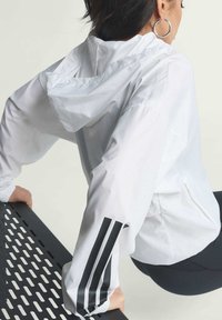 White lightweight jacket with a hood, featuring black three-stripe design on the left sleeve. Smooth texture, fitted style, and cinched cuffs.