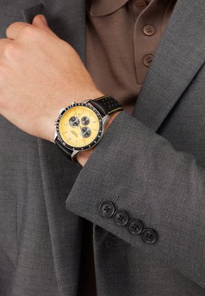 Black and yellow chronograph watch with a round case, leather strap, and silver hardware on a wrist, paired with a grey suit jacket.