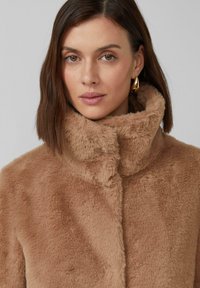Manteau en fausse fourrure marron clair avec un col montant. Texture douce et design sans coutures, avec une fermeture à l'avant et des accents minimalistes.