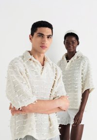Două modele poartă bluze tricotate supradimensionate, texturate, de culoare crem, cu un model țesut și detalii cu franjuri, asortate cu piese de jos deschise la culoare.