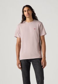 Lichtroze katoenen t-shirt met korte mouwen, ronde hals, en een klein rood Levi's logo op de linkerborst, gecombineerd met donkere jeans.