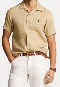 Camisa de lino beige de manga corta con un bolsillo en el pecho, botones en la parte delantera y un pequeño logo azul. Combinada con pantalones blancos y un cinturón trenzado marrón.