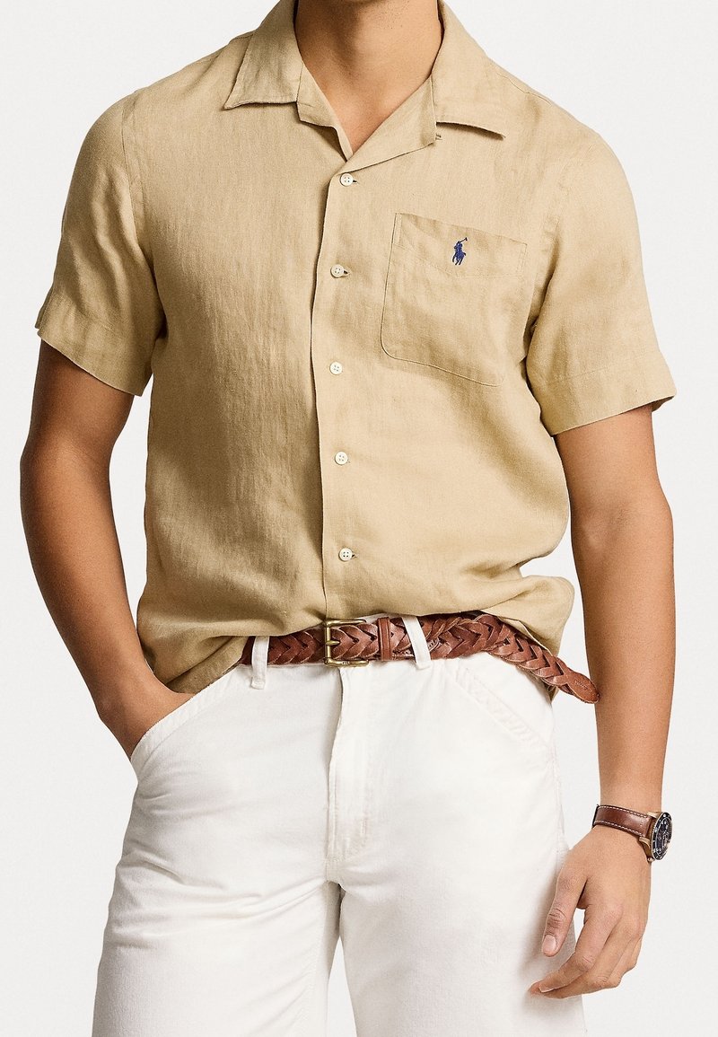 Camisa de lino beige de manga corta con un bolsillo en el pecho, botones en la parte delantera y un pequeño logo azul. Combinada con pantalones blancos y un cinturón trenzado marrón.