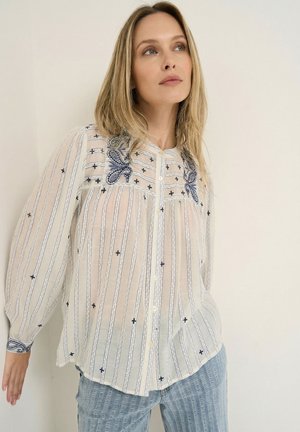 Frau trägt eine transparente weiße Bluse mit blauen Stickmustern und hellblaue strukturierte Hose, steht vor einer schlichten beigen Wand.
