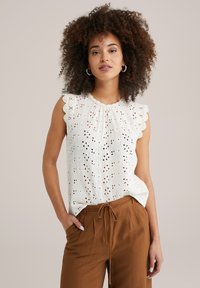 Witte mouwloze top met gekartelde randen, voorzien van oogjes uitsparingen; gecombineerd met bruine, ontspannen pasvorm broek en een trekkoord in de taille.