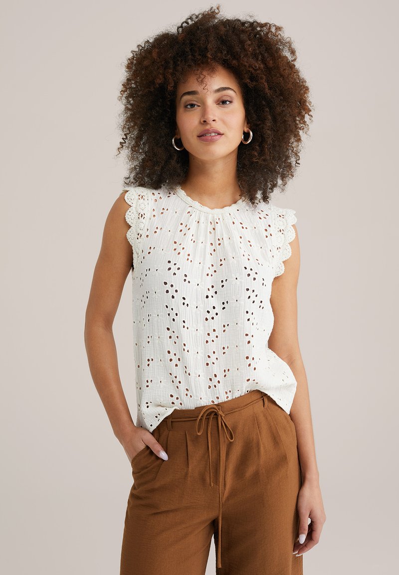 Witte mouwloze top met gekartelde randen, voorzien van oogjes uitsparingen; gecombineerd met bruine, ontspannen pasvorm broek en een trekkoord in de taille.