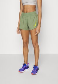 Pantalones cortos deportivos verdes con cintura elástica y un prominente logo de Nike en amarillo. Combinados con zapatillas de correr moradas con acentos en naranja.