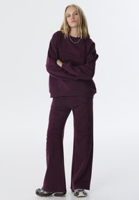 Burgunderfarbener Sweatshirt mit strukturierten Details, kombiniert mit passenden weiten Hosen mit bestickten Mustern. Das Modell steht in entspannter Pose.