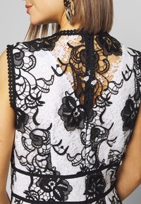 Biała sukienka z koronką, z czarną koronkową nakładką w kwiatowy wzór, ze skórkami i wycięciem w kształcie łezki z ukrytą zamkiem z tyłu. Czarny akcent w talii.