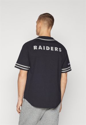 Mann trägt schwarzes Raiders-Trikot mit grauen Streifen an den Ärmeln, graue Sporthose, von hinten vor einfarbigem Hintergrund.