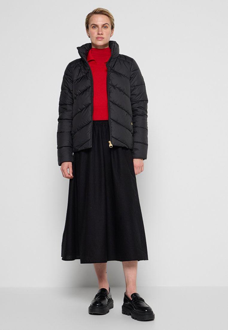 Schwarze Puffjacke mit Chevron-Muster und Stehkragen, getragen über einem roten Pullover und einem schwarzen Rock. Schwarze klobige Schuhe runden den Look ab.