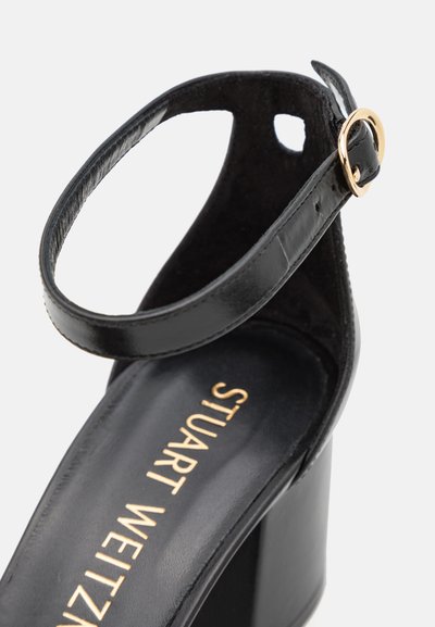 Sandale noire à talons avec un bout ouvert arrondi, dessus en cuir, une bride de cheville avec une boucle en or, et une semelle de plateforme texturée.