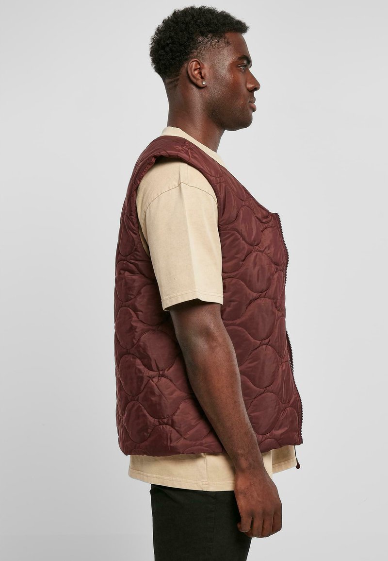 Gilet matelassé bordeaux avec fermeture éclair à l'avant, encolure arrondie et sans manches, porté sur une chemise beige, présenté en vue de profil.