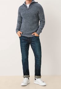 Grå stickad tröja med kvartslång dragkedja och ribbad textur, mörka denimjeans och vita sneakers med svarta detaljer.